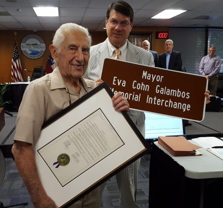 Dr. John Galambos (left) receives a proclamation and miniature sign from Mayor Rusty Paul recognizing the interchange naming at a June 7 City Hall ceremony. At rear, City Council members Tibby DeJulio and Andy Bauman look on, backed by a portrait of Eva Galambos. (Photo John Ruch)