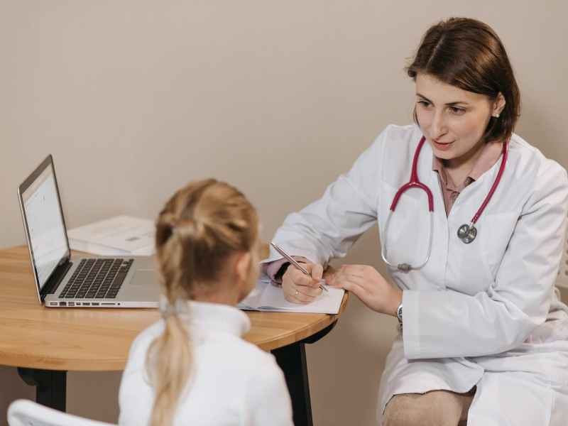 woman in white lab coat listening to a girl and writing down notes