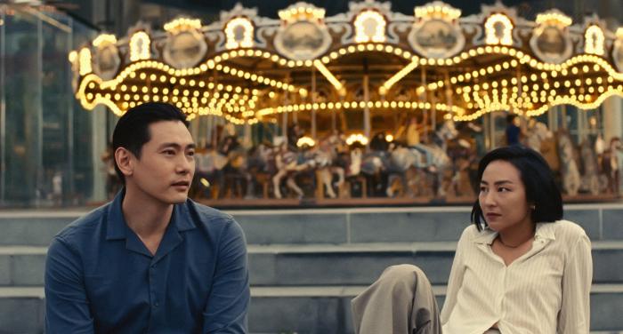 Teo Yoo and Greta Lee sit in front of a carousel in the movie "Past Lives."