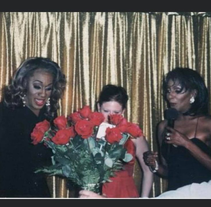 Tina Devore and Lady Chablis stand with director Nicole Sage in the middle. Nicole is partially obscured by red flowers and is wearing red. Lady Chablis and Tina Devore are wearing black. 